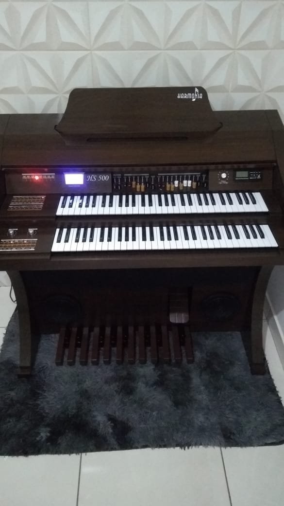 Órgão Eletrônico Harmonia HS-500 Luxo Marrom – 61 teclas no manual superior e 61 no inferior, pedaleira de 20 notas, drawbars completos, 160 sons prontos, Rotary Speaker, 4 memórias e Módulo USB/Bluetooth. Amplificação 120W RMS estéreo, conjunto de falantes de 10" e 8" + tweeters, gabinete 50mm e banqueta almofadada inclusa.